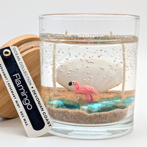 Candle in a glass jar with a flamingo figure and sand-like texture, on a white background.